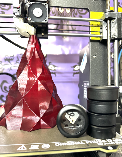 a red faceted Christmas tree printing on a Prusa MK4 3D printer with a stack of round, black containers belonging to Diamondback Nozzles with one facing forward so the logo can be seen on the top glass of the container.