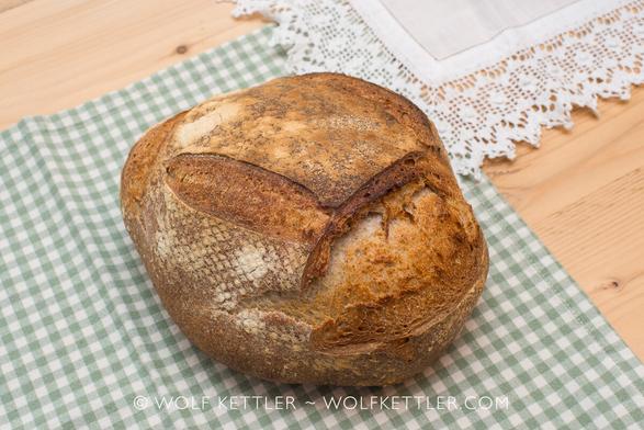 The photograph shows a crusty, rustic, golden loaf of bread on a green and white checked fabric placemat, seen from a high viewpoint.