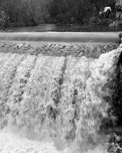 Monochromatic photo of a torrent of water flowing over a weir. The top section shows a placid river lined with trees, the surface broken by raindrops. A few people are walking alongside with umbrellas. The river then drops over a 3m high weir, so the lower half of the photo is just showing turbulent whitewater descending into a pool of foam at the bottom.