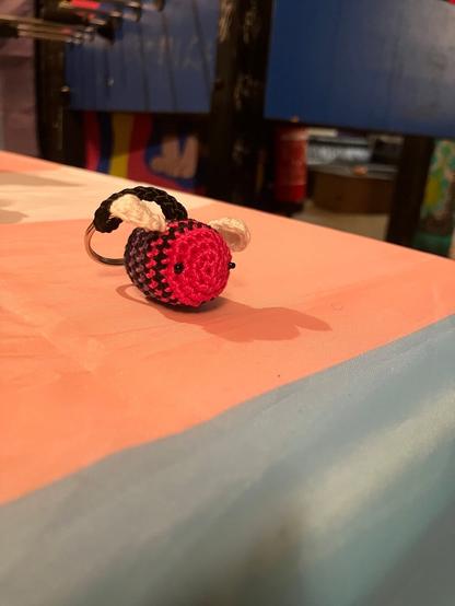 Man sieht eine von dino_versity gehäkelte Biene, die die Farben der Bi-Pride Fahne trägt. Also eine Bi-ene ;)
Sie liegt auf einem Tisch der mit einer Trans Pride Fahne abgedeckt ist.