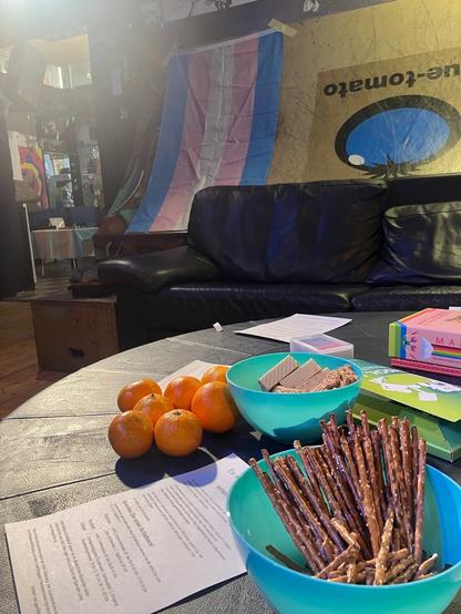 Man sieht einen Tisch mit Snacks und Info Material, im Hintergrund eine auf die Seite geschobene Skate-Ramp an der eine Trans Pride Fahne hängt.