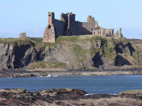 Tantallon Castle in East Lothian, seen from Seacliff. The image shows a rocky headland rising from the sea with the curving wall of Tantallon Castle cutting across it in the upper part of the frame, and more ruined stone buildings to its right. There are cliffs below the castle and sea this side of them, then rocks in the foreground. The sky is blue.