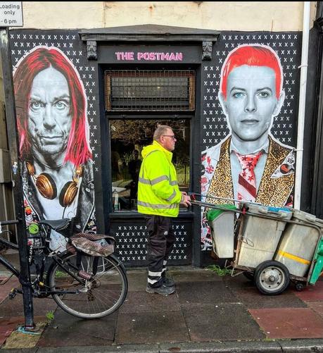 Streetart. On the outside wall of a store in Brighton, the two pop/punk singers Iggy Pop and David Bowie were sprayed/painted in pop art style around the black window. The faces are grey with colored elements. Framed by a black wall with countless white stars, David Bowie can be seen on the right with short red hair, a white shirt, a glittering gold jacket and a red tie with stars. He wears a pair of 80s buttons on his lapel. On the left is Iggy Pop in the same style with red hair, gold headphones around his neck, leather jacket and buttons. In the photo, a man from the city cleaning service in a neon yellow jacket pushes his car past David Bowie. A bicycle is chained to a pillar in front of Iggy Pop.
Info: "The Postman Art" are two anonymous street artists who collaborate with photographers to create artworks of icons past and present and give them a colorful punk twist. The colorful pasteups and murals can be seen throughout England and in museums around the world.The façade of the store is constantly being redesigned.