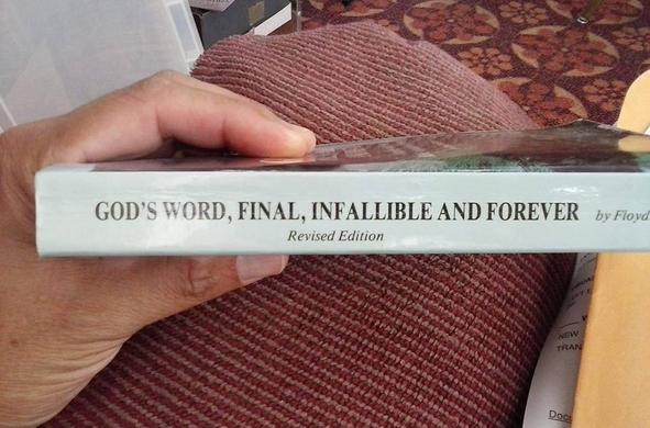 A person's hand holding up a book titled "God's Word, Final, Infallible and Forever" (by Floyd C. McElveen).
Below the title, in smaller text is the ironic "Revised Edition".
The image primarily shows the spine. The background could be described as "busy", with parts of furniture, carpeting and a wall visible.