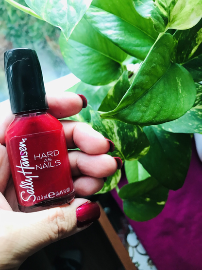 Author holding red nail polish, showing freshly painted long red nails in front of green leaves house plant