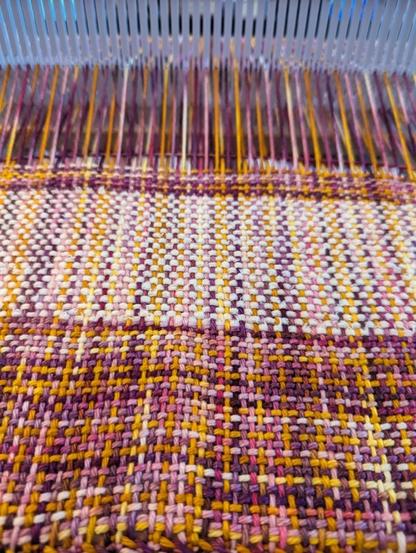 A piece of woven cloth being made on a rigid heddle loom. It features a variegated pink/purple/orange colour and a lightly fluffy white woven in wide stripes.