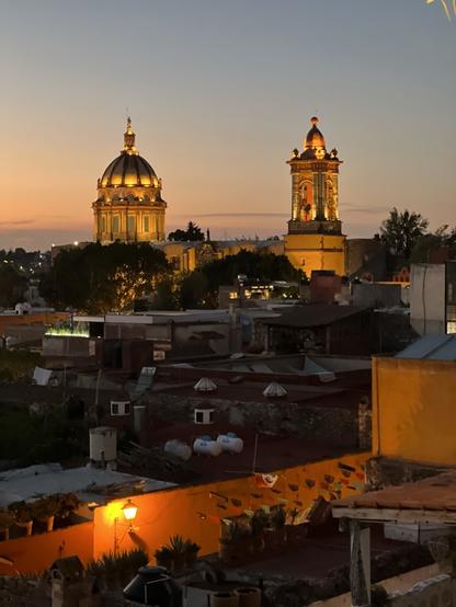 Lit up cathedral towers in the distance. Rooftops of homes and businesses can be seen in the foreground shortly before sunset.