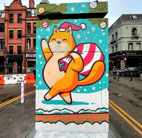 Streetartwall. The mural of a cheerful cat has been painted/sprayed in cartoon style on a small, narrow stand in a public square. The brown/white cat with a red cap is trudging upright and waving through the falling snow. She is carrying a red and white striped sack on her back.