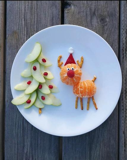 Photograph. A color photograph of a white plate with a Christmas scene of food and a "cute" reindeer. The reindeer is standing next to a Christmas tree: Rudolph's head is made from a tangerine, his body from half a tangerine (so that he lies flat on the plate). His triangular hat is made from a piece of strawberry with a mini pom-pom made from a white marshmallow. His legs, tail and antlers are made from brown pretzels. The nose is a raspberry and the eyes are tiny white edible candy eyes. (Marshmallow or cream with chocolate pearls also works) The Christmas tree next to it is made from wafer-thin green apple slices and decorated with red cranberries