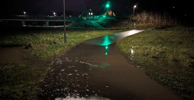 Ein auf einer Strecke von einigen Metern unter Wasser stehender Radweg durch eine in einer Mulde gelegenen Wiese.