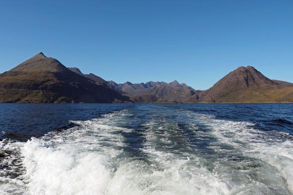 Blick von einem Boot auf eine dunkle Bergkette vor strahlend blauem Himmel, das Kielwasser ist weiß verwirbelt. 

View from a boat of a dark mountain range against a clear blue sky, the wake swirled with white.