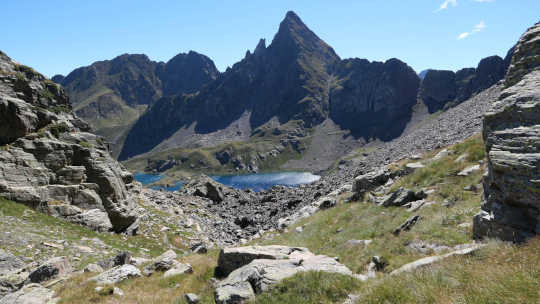 Paysage de montagne avec au premier plan une pente herbeuse parsemée de rochers, en s'éloignant la pente descend en devenant plus minérale. Au loin, des lacs turquoises reflètent le Soleil. Ils sont surplombés par une ligne de crête escarpée et ombragée. Le ciel est clair.