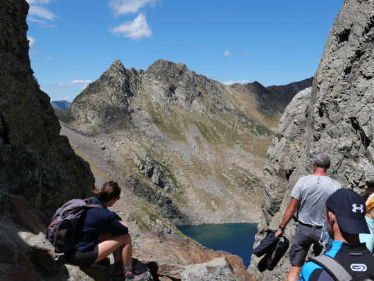 Vue depuis le port de Vénasque. Au premier plan, des randonneurs : dos tournés marchant vers la France ou assis de profil regardant en contrebas, le sac encore sur le dos. Entre deux pans rocheux, le paysage est constitué d'un ciel clair pourvu de rares nuages blancs et d'une montagne rocheuse qui domine un bout de lac bleu profond,