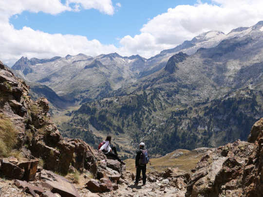 Vue depuis le port de Vénasque vers l'Espagne. Dans un premier plan rocheux, légèrement teinté de couleur rouille, deux randonneurs équipés contemplent le grandiose panorama sur la Maladeta, dont une partie seulement apparaît sur la photographie. L'un est debout, l'autre assis appuyé sur un bras tendu. Sous un ciel à moitié nuageux, l'arête altière et sombre n'est qu'un liseré qui surplombe comme un immense ébouli gris de plus en plus piqué du vert foncé des arbres et du vert pâle de l'herbe. La neige et la glace assurent encore une présence minimale sous les sommets.
