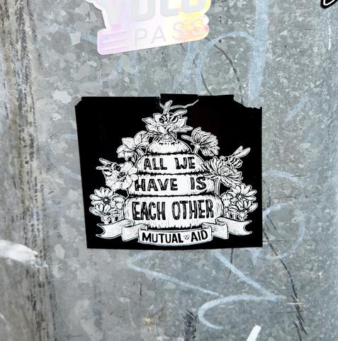 A black and white sticker on a steel lamp post in Baltimore. A N.O. Bonzo design stating All We Have Is Each Other - Mutual Aid