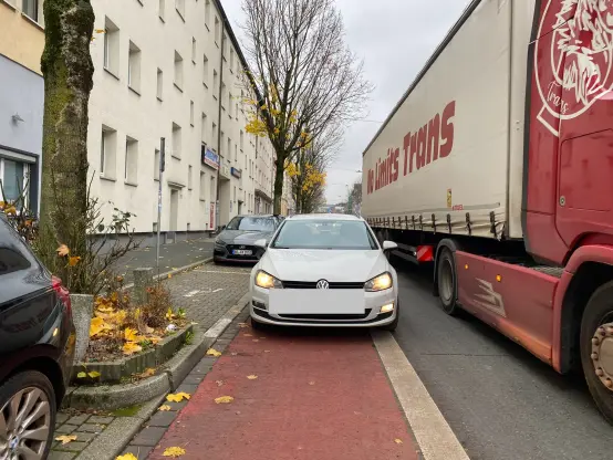 Ein Auto parkt auf einem roten Radweg direkt neben einem freien Behindertenparkplatz. Neben dem Auto fährt ein LKW