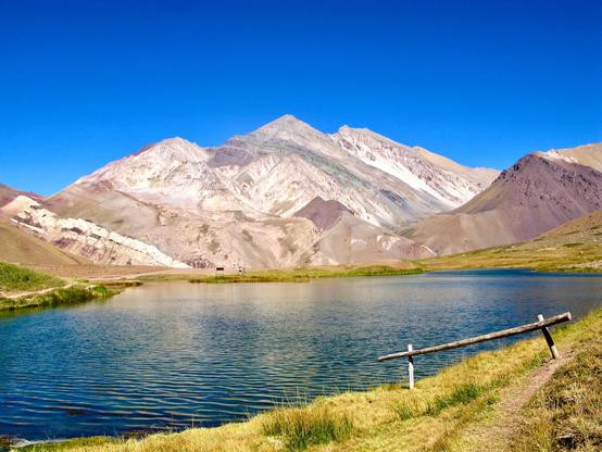 An high mountains body of water occupies the lower half of the image. In the background stands a three peaked mountain exposing reddish and whitish interesting geological bands. Sky is cloudless and deep blue.