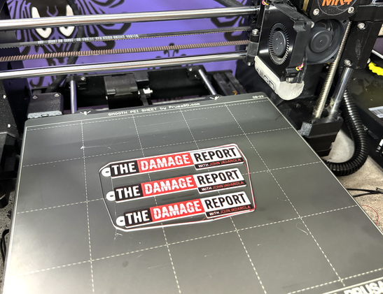 3D printed keychains depicting The Damage Report With John Iadarola logo in the appropriate colors, black, white and red. They are still on the bed of the Prusa MK4 they were printed on.
