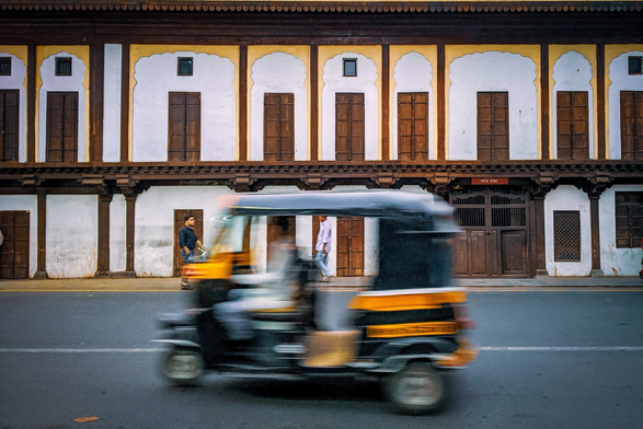 Photo of an auto rickshaw in blurred motion driving along a road with traditional Indian buildings behind