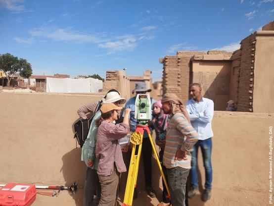 Foto mehrerer Personen in praktischer Feldbekleidung, die vor Lehmziegelarchitektur um eine Totalstation gruppiert sind.