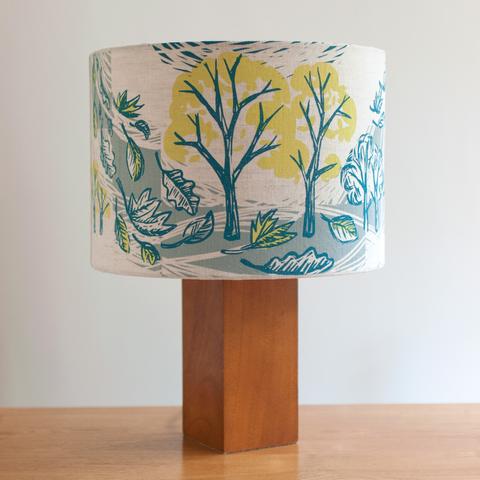 A hand printed lampshade with trees, blowing leaves and a blue background. The trees are in dark blue and yellow. The leaves are in dark blue, white and yellow. The background is abstract shapes.