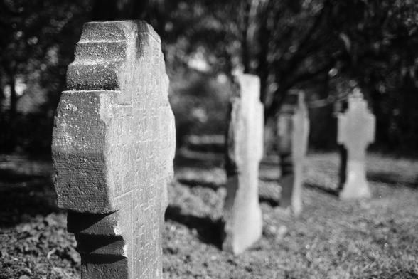 Analoges schwarz-weiß Foto: Vier Grabsteine in Form von Kreuzen stehen in einer Reihe. Nur das vordere ist scharf, die anderen werden mit zunehmender Entfernung unschärfer. Im Hintergrund unscharfes und von der Offenblende verzerrtes Gebüsch.