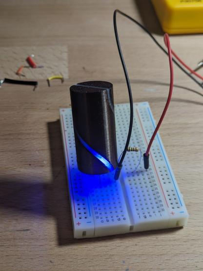 A cylindrical 3D printed object is on a electrical breadboard. There is a helical section of the cylinder that is glowing blue at the bottom of the form. Wires are connected to the breadboard.