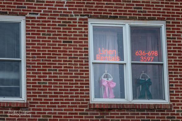 A white framed windows in a brick building contains 2 red neon signs reading "Linen Rentals" and "636-698-3597" along some linens on hangers.