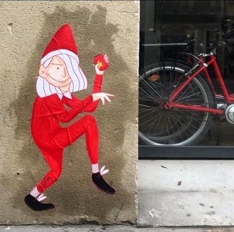 Streetart. In the bottom corner of a beige wall next to a store is a small sticker with a drawing of a girl with pink hair, dressed like an elf in a tight red suit and a Christmas hat.  She is holding a tin can with a golden star on it and is walking carefully towards the bike store with a smile. She probably wants to give herself a present.