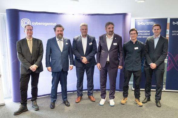 Das Bild zeigt eine Gruppe von sechs Männern für ein Foto. Sie stehen vor einem Hintergrund der Cyberagentur sowie der Brandenburgischen Technischen Universität Cottbus-Senftenberg. Die Männer tragen geschäftliche Kleidung – Anzüge und Hemden, einer von ihnen trägt eine Krawatte. Ihre Kleidungsstile variieren etwas, mit unterschiedlichen Farben und Schnitten von Anzügen und Schuhen. Das Fotos entstand während der Vertragsunterzeichnung für Projekt der Cyberagentur mit Zander Laboratories. 
Foto: Britta Zwigart/Cyberagentur