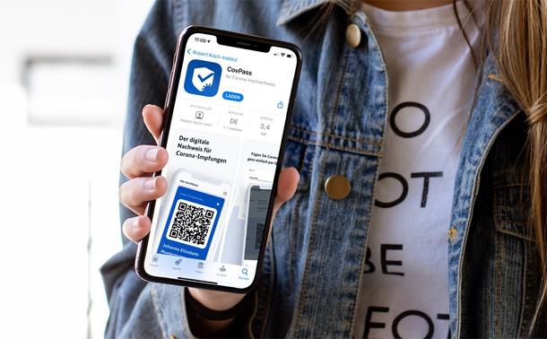 Eine Person hält ein iPhone in die Kamera, auf dem die CovPass-Produktseite des App Stores geöffnet ist.