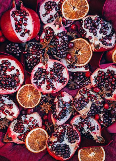 A horizontal image of a pomegranate and preserves spread on a burgundy cheesecloth background, photographed from above. At random in the frame there are preserved orange slices, cranberries, pomegranates and loose arils, matching burgundy ribbon, and whole star anise.