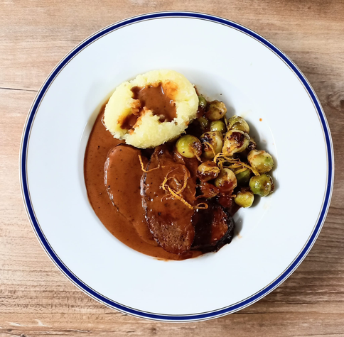 Auf einem Teller zwei Scheiben veganer Sauerbraten mit Sauce, Rosenkohl und einem grünen Kloß. Rosenkohl und Braten sind mit Orangenzesten bestreut.