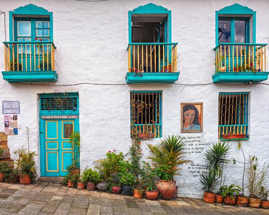 A two story white washed building on a sloping street has three second story balcony windows and two first story windows with turquoise trim. There is a turquoise colored door and flower pots with various sized plants along the ground.