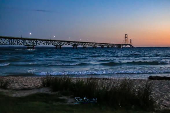Mackinac Bridge at Sunrise. Low light, pink sky on the horizon and blue sky above. Blue water with small waves. Some lights on the Mackinac Bridge. Brown sand and beach grass in the foreground.  Image at:  https://beautifulsunphotography.com/featured/mackinac-sunrise-deb-beausoleil.html  See more art & blog at: https://beautifulsunphotography.com/ https://debbeautifulsunphotography.com/  https://debbeausoleil.com