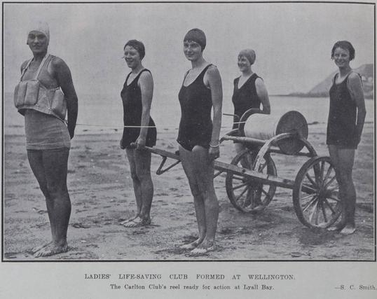 Black-and-white group photo: Ladies Life-saving Club Formed at Wellington. The Carlton Club’s reel ready for action at Lyall Bay, Wellington. Description: Five women in swimming togs, three wearing swimming caps, stand on a beach next to a two-wheeled life-saving reel. A bay and hill by the coast can be seen in the background. Citation: Auckland Weekly News, 6 January 1927, p. 14. Auckland Libraries Heritage Collections AWNS-19270106-14-02. https://kura.aucklandlibraries.govt.nz/digital/collection/photos/id/259073