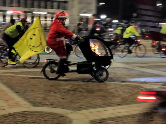 Einige Menschen auf Fahrrädern fahren durchs Bild. Im Vordergrund ein als Weihnachtsmann verkleideter Fahrer mit weihnachtlich geschmückten Lastendreirad