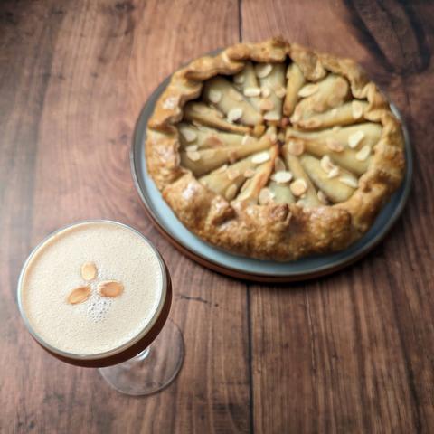 A brown cocktail in a Nick and Nora garnished with three small toasted almonds on top, sitting in front of a galette pie containing parsnips and topped with toasted almonds. They are both sitting on a brown wood effect background.
