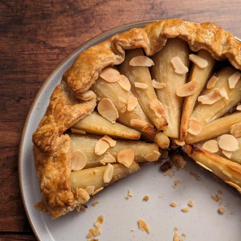 A galette pie containing parsnips and topped with toasted almonds, sitting on a plate on a brown wood effect background. A large slice has been cut from the galette, and some crumbs are spread on the plate.