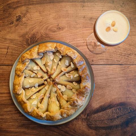 A galette pie containing parsnips and topped with toasted almonds, next to a brown cocktail in a Nick and Nora garnished with three small toasted almonds on top. They are both sitting on a brown wood effect background.