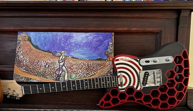 A 3D Printed Guitar with a wooden neck and metal hardware. Black core with red hexagons. Sitting on an old, beat up piano.