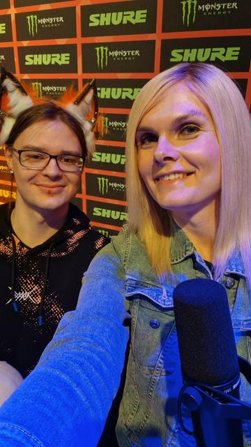 Lena rechts und J0J0 links vor einer Werbewand. Beide lächeln.
