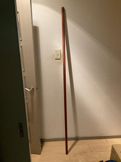 A 182cm long wood staff, walnut color, leaning on a white wall in a house.