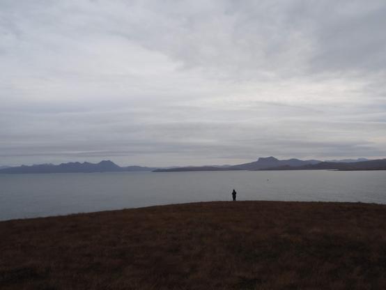 looking across a sea loch, hills of Assynt and Coigach on the horizon, cloudy sky. In the foreground a small figure stands silhouetted against the sea