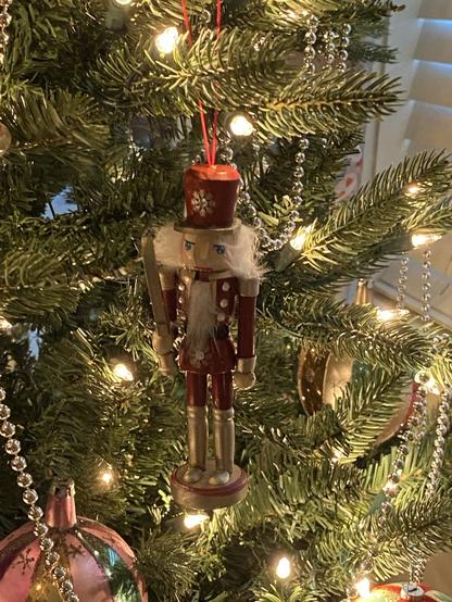 Newly acquired Nutcracker ornament hanging on a branch of our Christmas tree. He is dressed in a red hat, military style jacket, and pants, with tall golden boots. He is grasping his sword in his hand - ready to do battle with the mouse king. He is surrounded by warm white tree lights and there is some silver bead garland and large glass ornaments on the edge of either side of the frame.