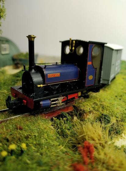 Photo of a 1:76 model of blue narrow gauge steam engine riding over a small bridge on a model railway layout with grass and yellow and red flower imitations at the foreground.