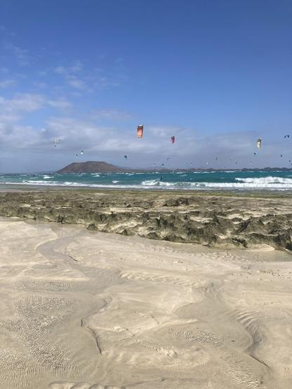 A beach view towards a small island surrounded by an ocean that has many waves due to it being very windy. There are many many windsurfers in the water. In the foreground there are some rocks and the sandy beach. The sky is blue with some clouds on the horizon.