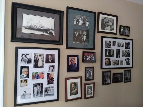 A light brown wall displays many framed photos of ancestors.  One photo on the top left displays a ship that carried ancestors to Ellis Island, New York, USA.  Another photo on the top left portion of the wall shows an old white-haired woman seated near a large spinning wheel.