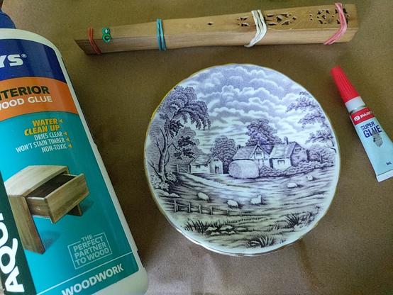 On a brown paper surface sit a teacup saucer painted with a rural scene in lavender and white, a carved wooden fan held closed by four rubber bands, a big bottle of wood glue, and a small tube of superglue.