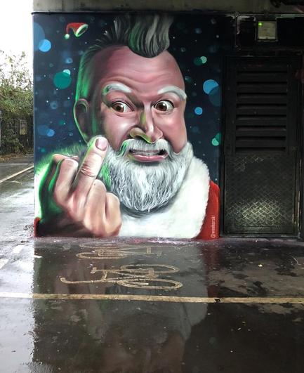 Streetartwall. The mural of a punky Santa Claus was sprayed on a concrete wall next to a parking lot. The background is blue with green and blue circles. In front of it is a Santa Claus with a punk hairstyle and white beard, giving us the middle finger. A little Santa hat flies away.